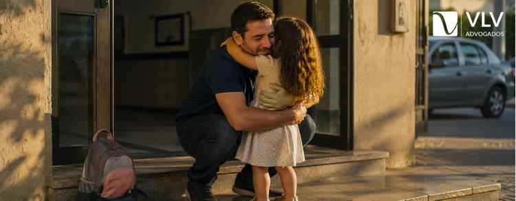 Pai abraçando a filha na entrada de um prédio residencial.